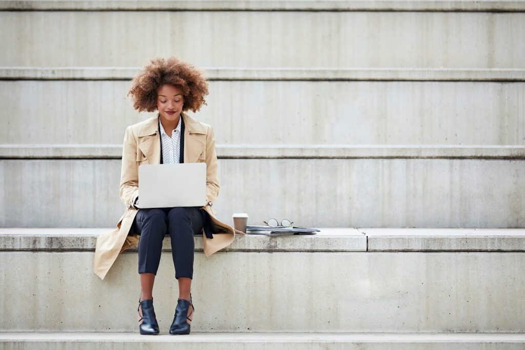 A woman, seated outdoors on a concrete grandstand, smiles and works at a laptop computer; beside her are a cup of coffee, a pair of eyeglasses, and some folders full of documents.