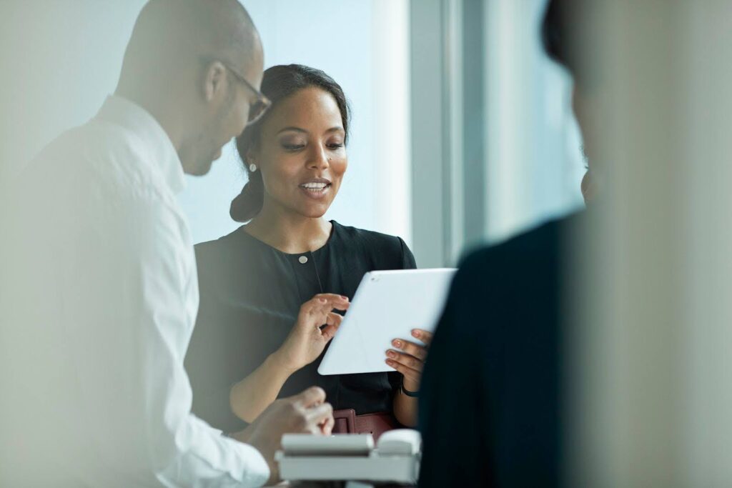 A woman in business attire enters information into a tablet while conversing with a professional associate.