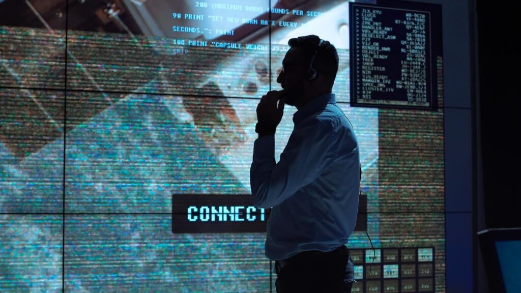 A man stands in front of a wall of large-screen monitors displaying data and text.