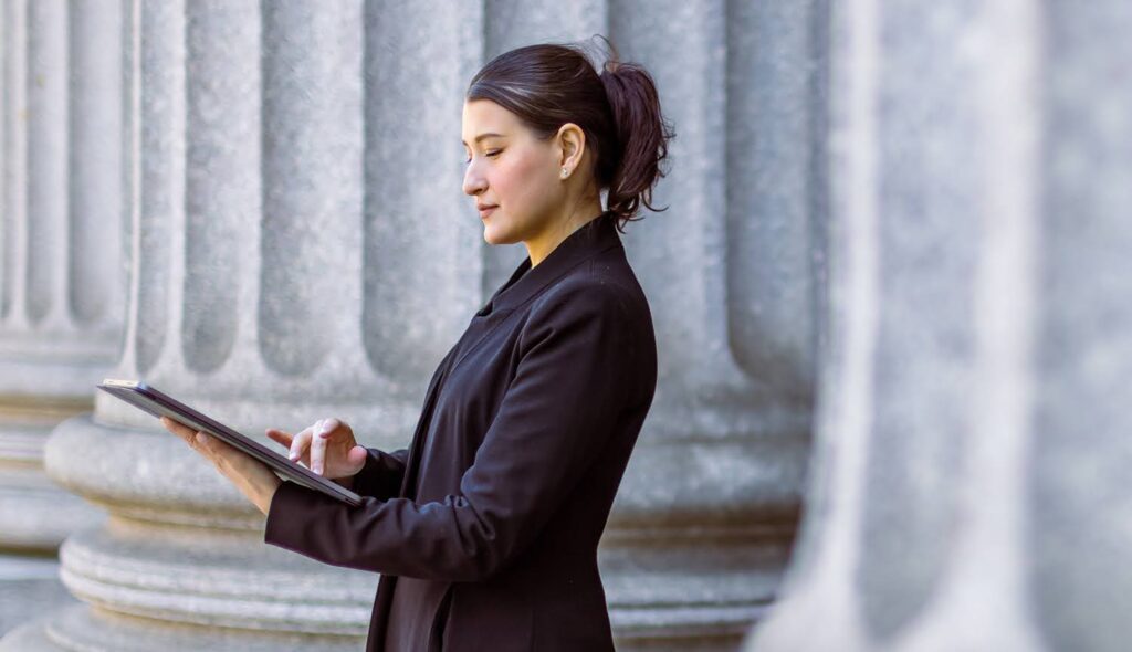 A woman in professional attire stands outside a courthouse building; she is using a tablet computer.