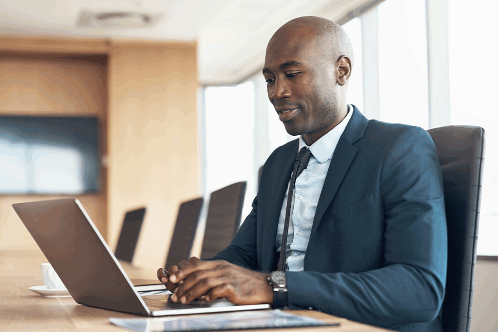 A focused businessman in a suit sits at a conference table in a modern office setting; he is typing on a laptop with documents nearby.