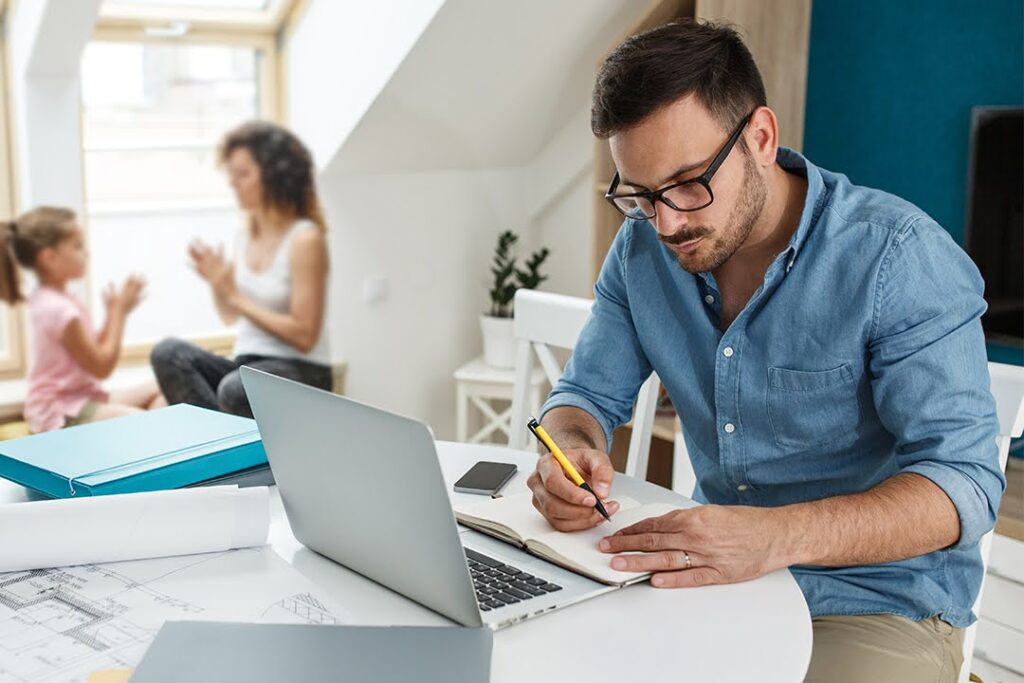 An adult seated at a table works at a laptop from home; their partner and child play in the background.