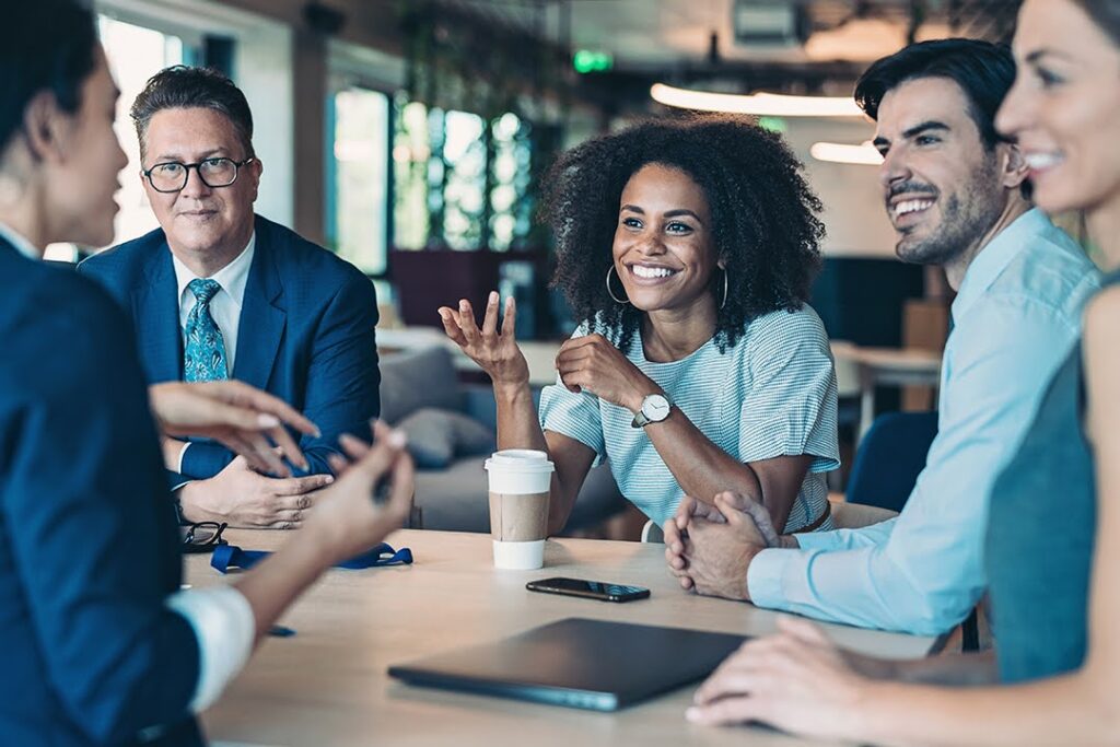 A diverse group of professionals sitting at a table, smiling and engaged in a lively discussion in a modern office setting.