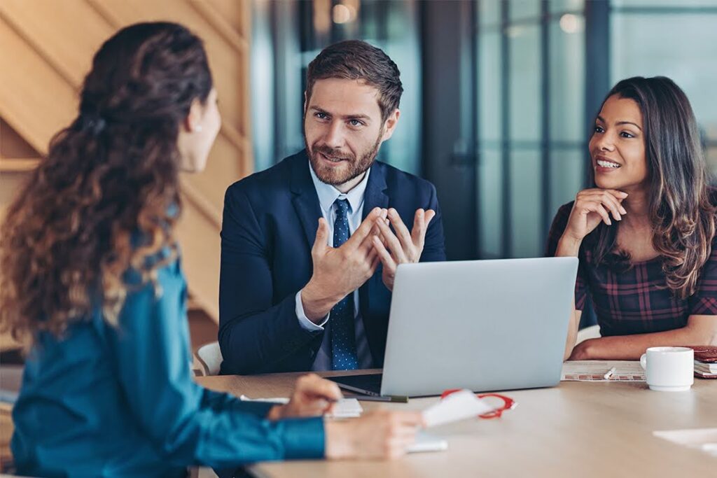 Three colleagues seated at a table in a business environment converse.