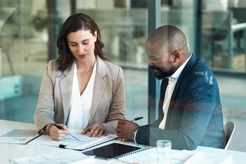 Two business professionals sit at a desk reviewing documents together, smiling as they converse in a modern office setting.