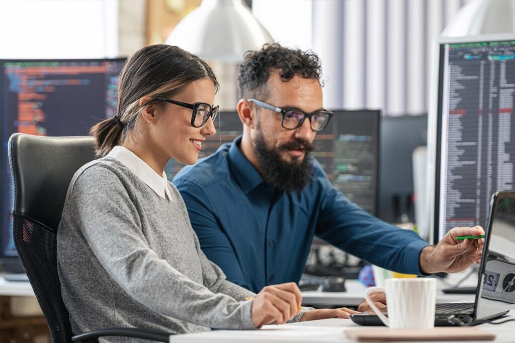 Two professionals seated at a desk with multiple monitors displaying code collaborate.