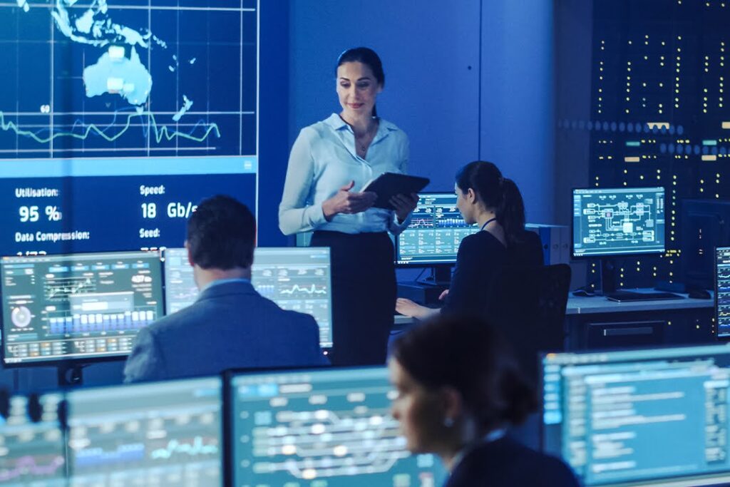 Professionals work at computer stations in a high-tech control room, as a woman holding a tablet stands and speaks, with data visualizations on large screens.