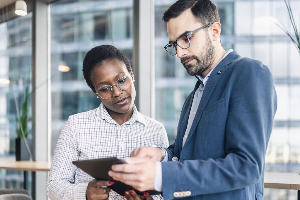 Two professionals standing in a modern office review information on a tablet.