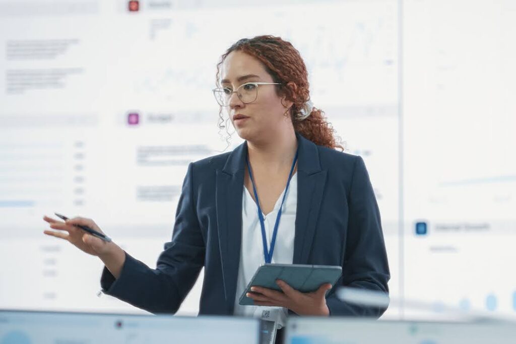 A woman in business attire holding a tablet and pen speaks in front of a large screen displaying data charts and graphs.